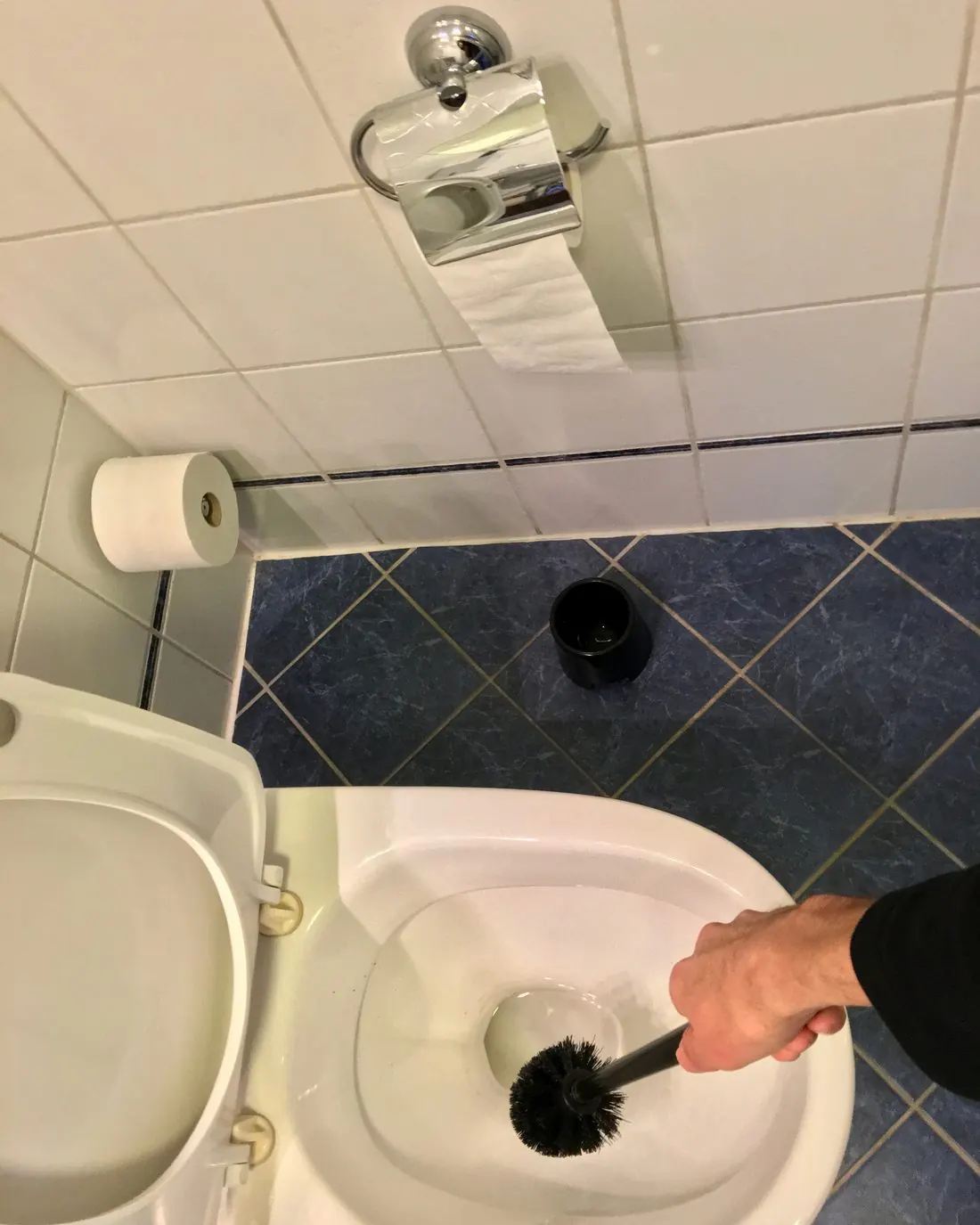 Mano limpiando la taza de un inodoro con escobilla en un baño alicatado de azul